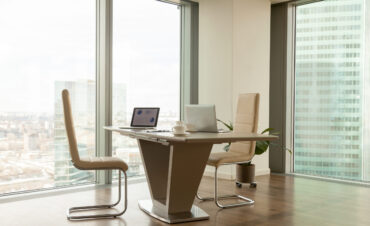 Image of work desk with two laptops on it and comfortable armchairs beside in bright office interior with large windows and urban landscape outside. Modern office workplace, negotiation room design